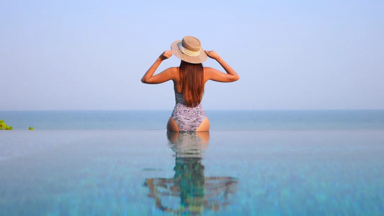 Back of a woman in swimwear sitting on infinity pool border touching her sun hat and raising hands up with a stunning view of tropical sea horizon in Miami Florida static