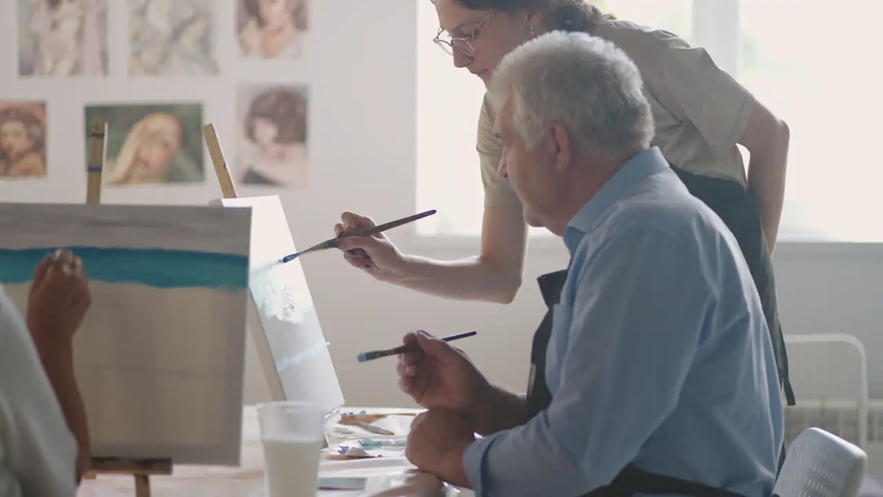 A female teacher shows a retired man how to draw a picture with paints and a brush at courses for the elderly A senior man draws a picture to a group of pensioners