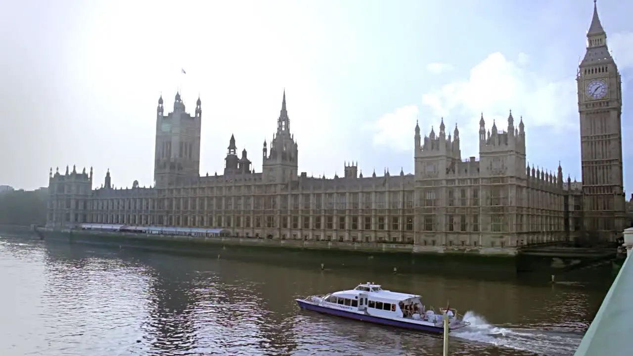 Houses of parliament with a long tourist boat going through the river thames on a very sunny day in london england