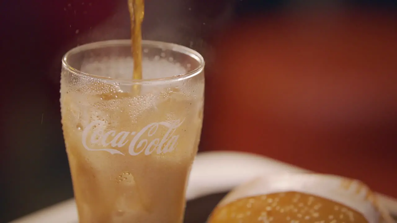 Pouring Coke On An Empty Glass With Ice And Burger Beside It