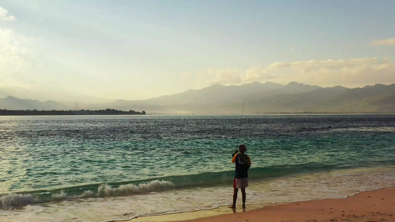 Young man reeling in his line while fishing in the shallow waves of an exotic beach during the early evening