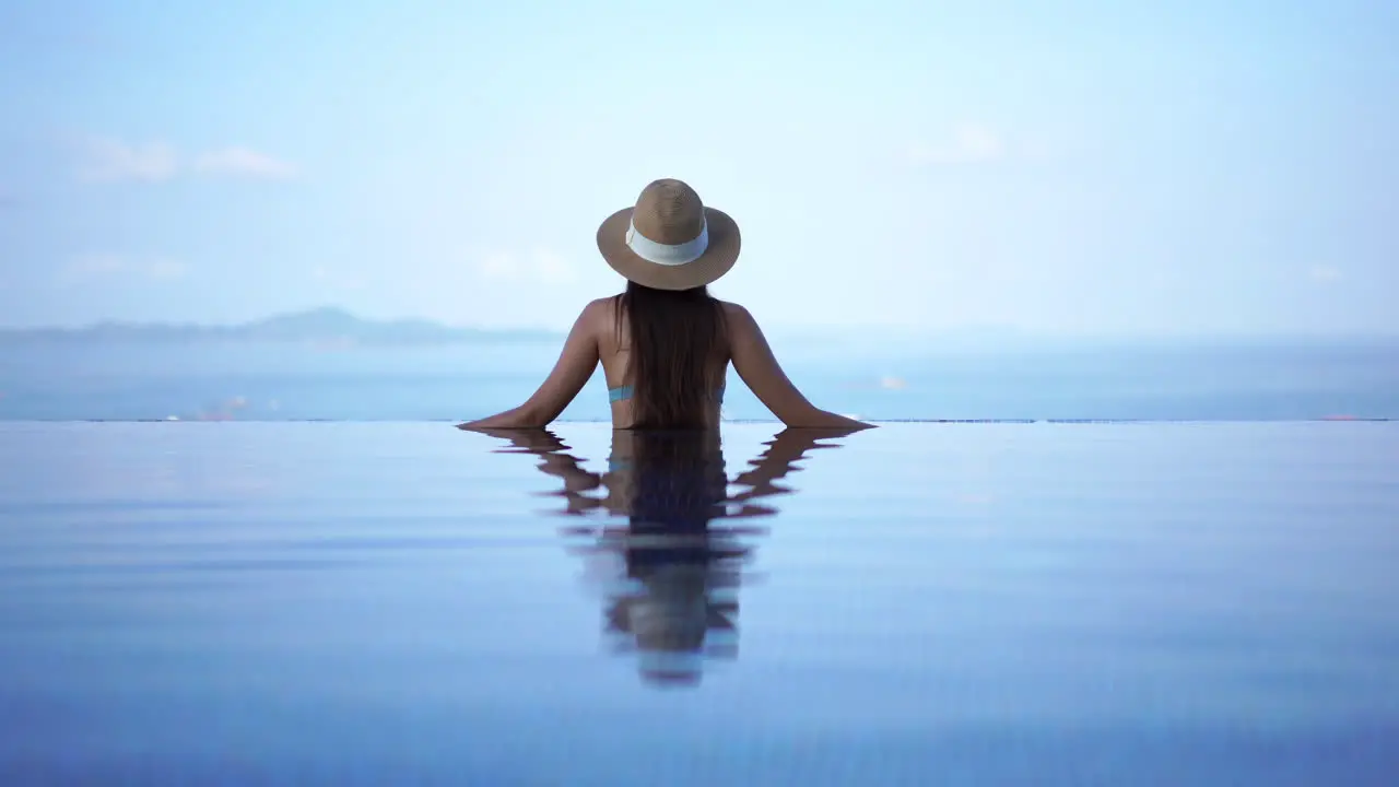 Back and Water Reflection of Female in Swimsuit and Hat Standing at Border of Infinity Pool and Looking at Horizon