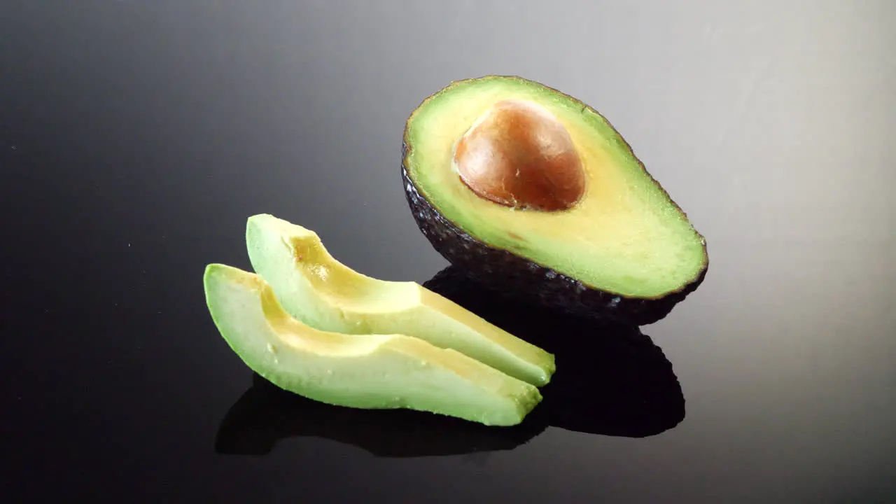 A ripe avocado cut in half with the pit and two slices ready isolated on a black mirror background