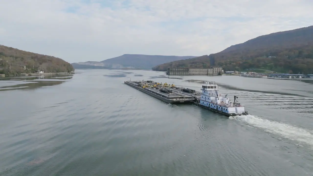 Aerial footage of a boat pushing a large industrial barge through the Tennessee River past Historic Hales Dam Powerhouse in Autumn