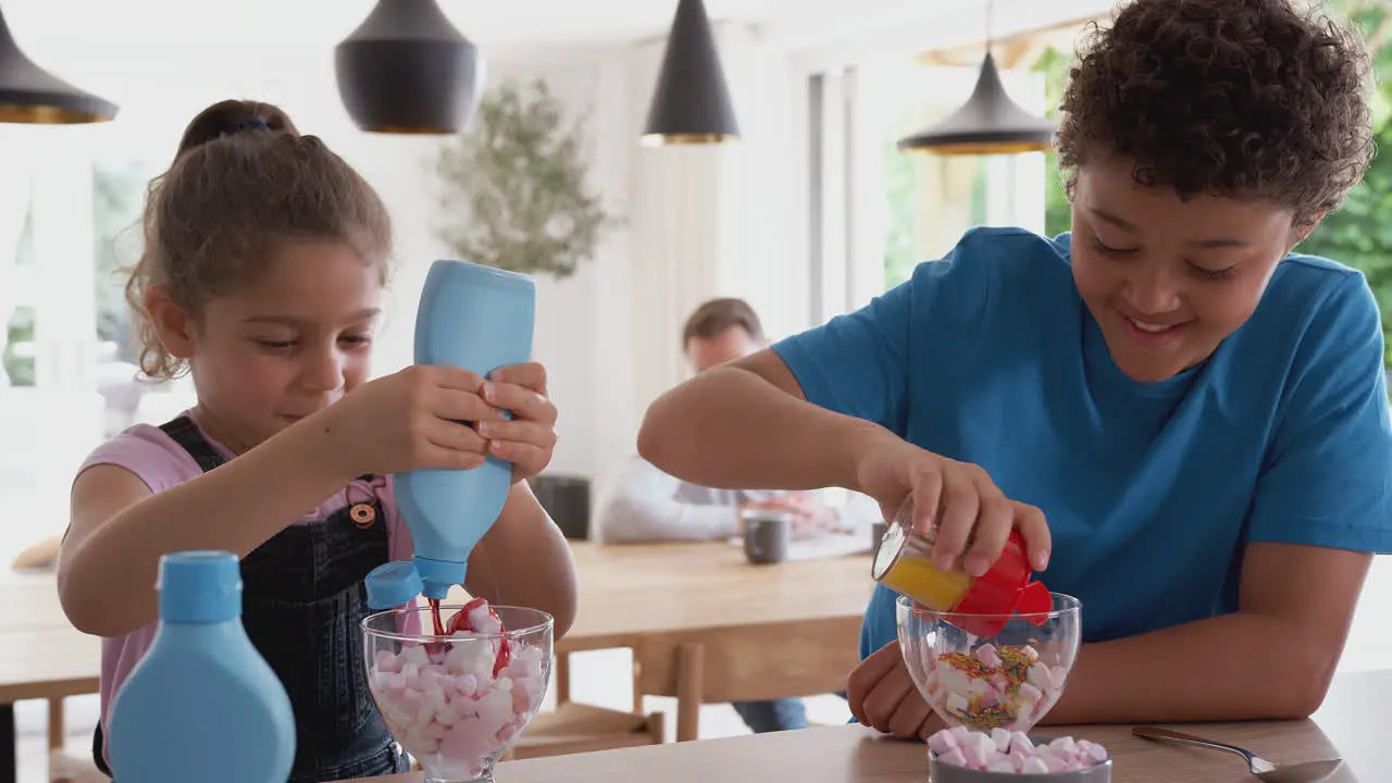 Children In Kitchen At Home Adding Sprinkles And Sauce To Ice Cream Dessert