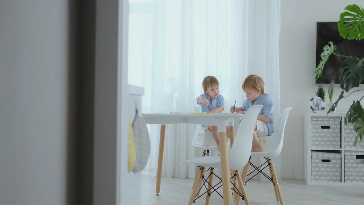 Two boys brothers sit at a table in the living room and draw with pencils on paper Mom comes up and hugs the boys smiling and stroking his head Praises and helps to make the task