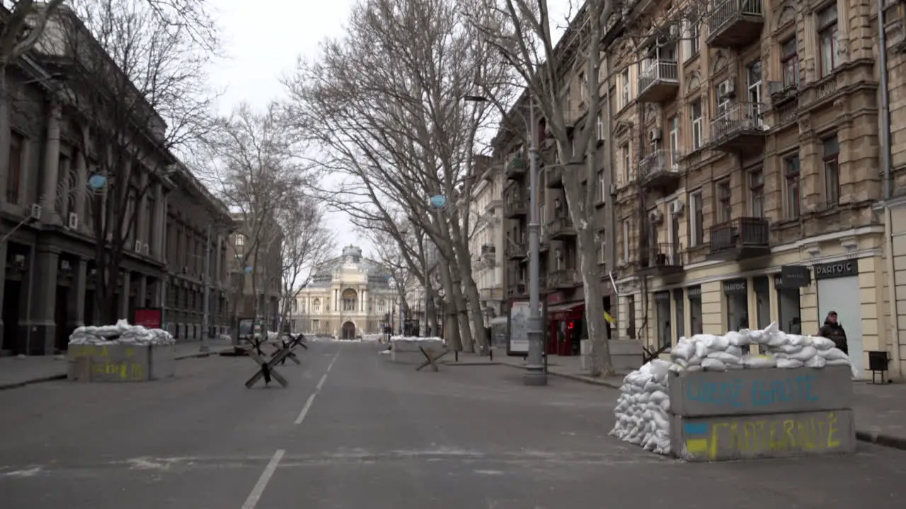 People walk past concrete barricades one of which is sprayed painted in yellow and blue spray paint with the words Liberté égalité fraternité” in front of the Odesa Opera and Ballet theatre