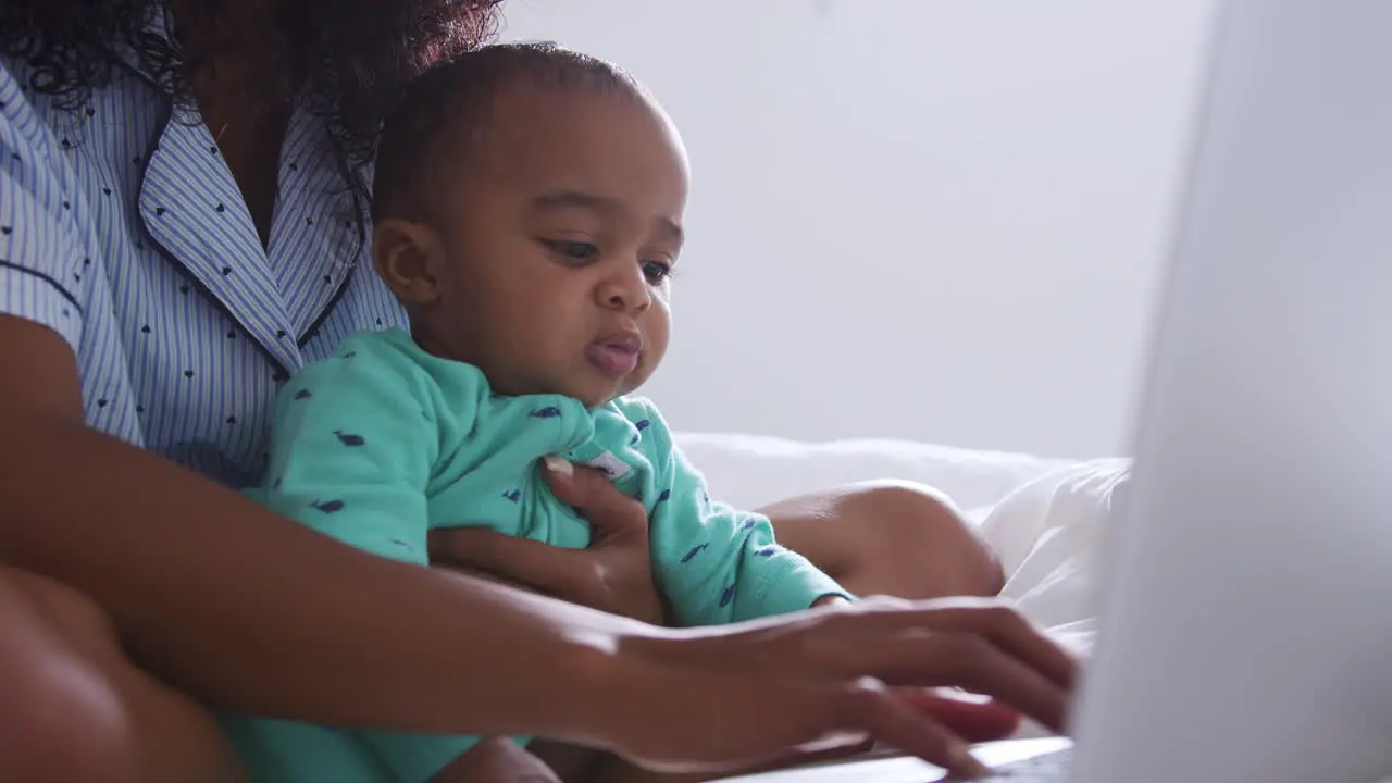 Mother Wearing Pyjamas Cuddling Baby Son In Bedroom Whilst Working From Home On Laptop