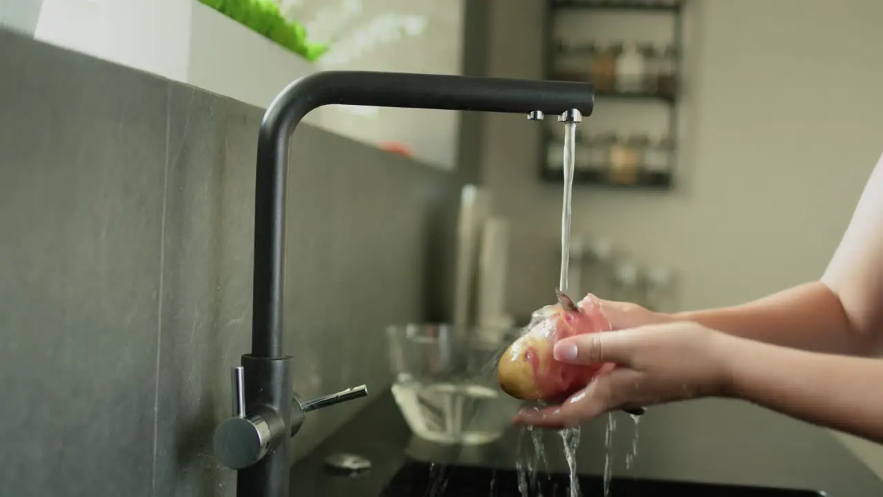 A woman peels potatoes under running water from a kitchen faucet