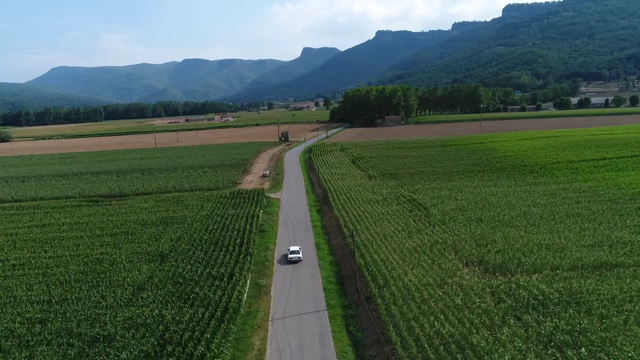 White Classic BMW Car Travelling Close To Hostalets de Bas In Olot Catalonia Spain Passing By Wide Green Fields Background With Foggy Mountains Under The Cloudy Sky Aerial Shot