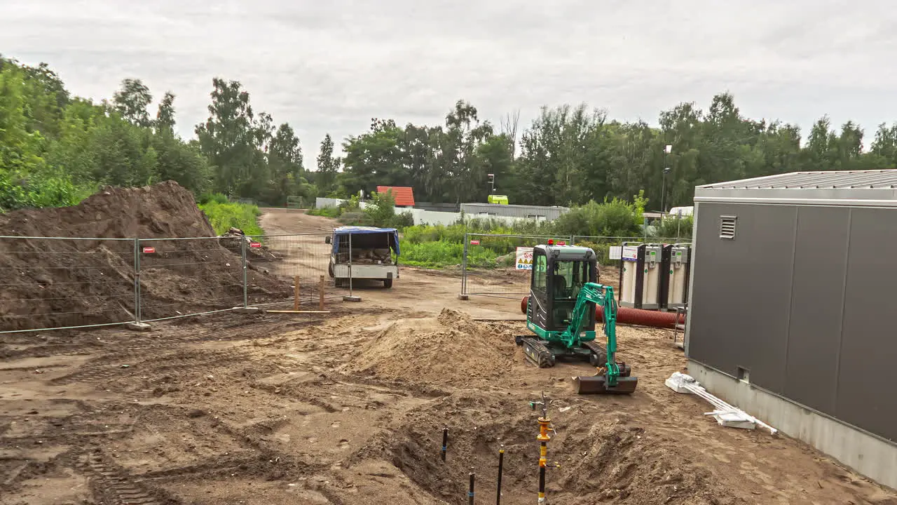  High angle shot of green excavator leveling earth for construction of a new parking lot beside newly made godown on a cloudy day in timelapse