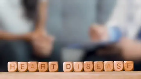 Medical Concept With Wooden Letter Cubes Or Dice Spelling Heart Disease Against Background Of Doctor Talking To Patient