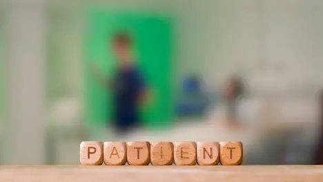 Medical Concept With Wooden Letter Cubes Or Dice Spelling Patient Against Background Of Nurse Talking To Patient In Hospital Bed