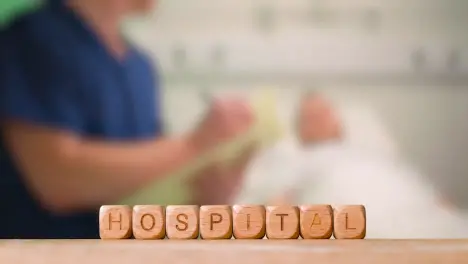 Medical Concept With Wooden Letter Cubes Or Dice Spelling Hospital Against Background Of Nurse Talking To Patient In Hospital Bed