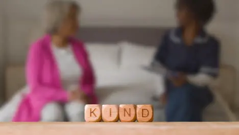 Medical Concept With Wooden Letter Cubes Or Dice Spelling Kind Against Background Of Nurse Talking To Patient In Hospital