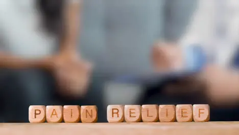 Medical Concept With Wooden Letter Cubes Or Dice Spelling Pain Relief Against Background Of Doctor Talking To Patient