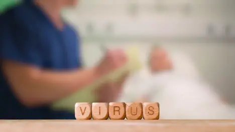 Medical Concept With Wooden Letter Cubes Or Dice Spelling Virus Against Background Of Nurse Talking To Patient In Hospital Bed