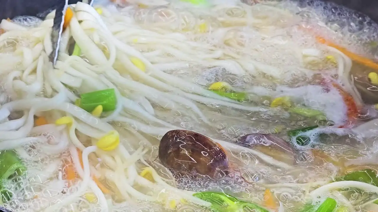 Seafood Clam Kalguksu Boiling in Pot Tongs Mix Knife Cut Noodle Soup While Cooking closeup