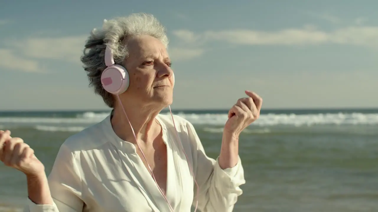 cinematic video of an elderly woman dancing on the beach near the water while wearing headphones and playing music