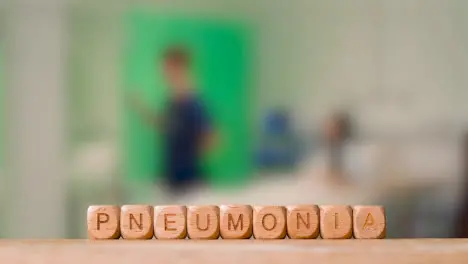 Medical Concept With Wooden Letter Cubes Or Dice Spelling Pneumonia Against Background Of Nurse Talking To Patient In Hospital Bed