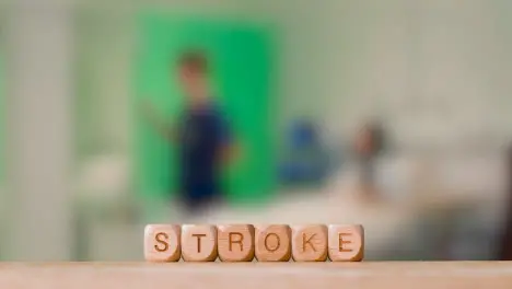 Medical Concept With Wooden Letter Cubes Or Dice Spelling Stroke Against Background Of Nurse Talking To Patient In Hospital Bed