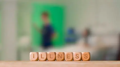 Medical Concept With Wooden Letter Cubes Or Dice Spelling Illness Against Background Of Nurse Talking To Patient In Hospital Bed