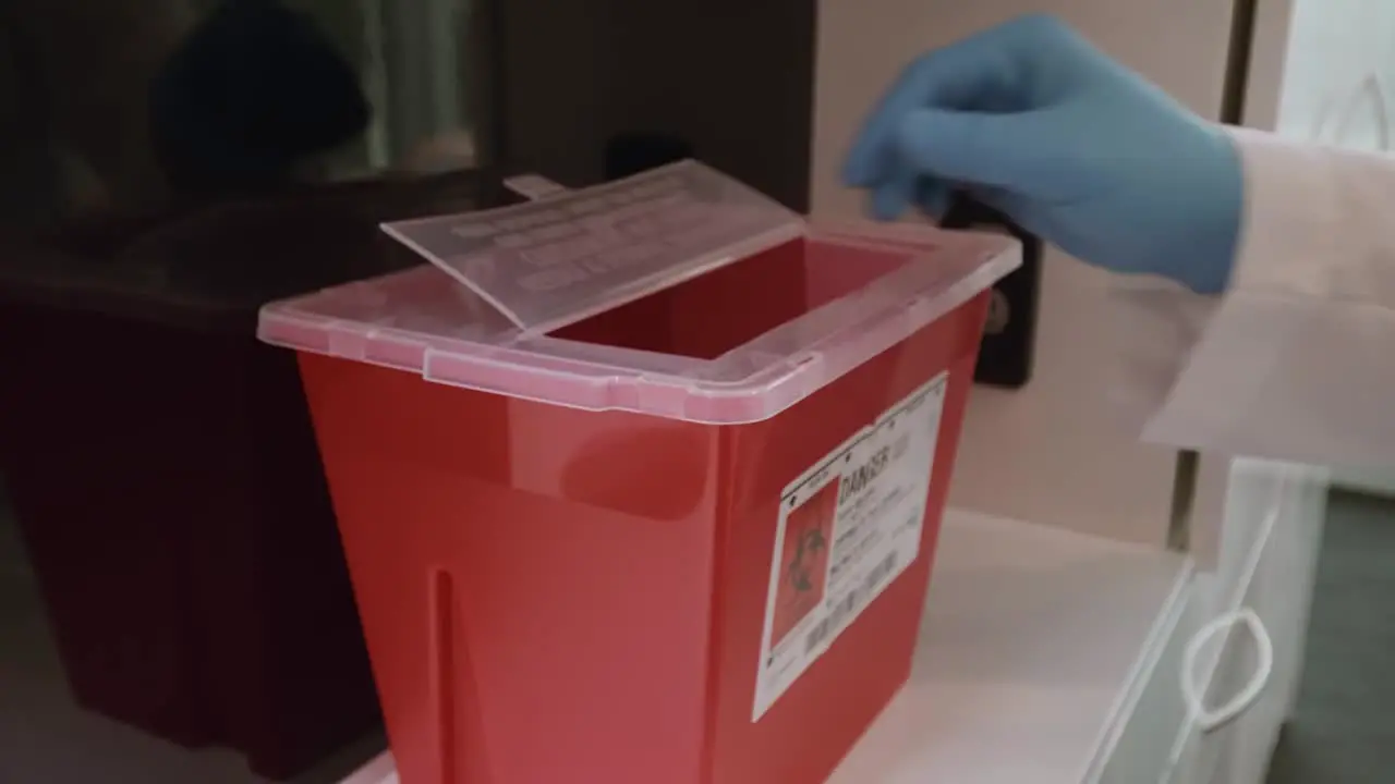 Gloved hand throwing empty vial and contaminated needle in sharps disposal container