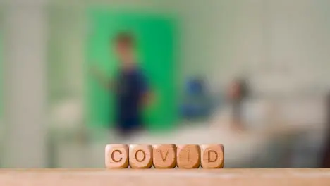 Medical Concept With Wooden Letter Cubes Or Dice Spelling Covid Against Background Of Nurse Talking To Patient In Hospital Bed