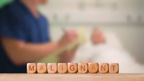 Medical Concept With Wooden Letter Cubes Or Dice Spelling Malignant Against Background Of Nurse Talking To Patient In Hospital Bed