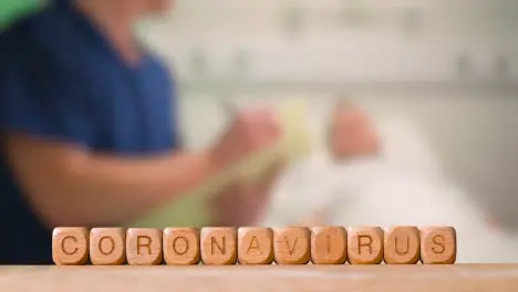 Medical Concept With Wooden Letter Cubes Or Dice Spelling Coronavirus Against Background Of Nurse Talking To Patient In Hospital Bed