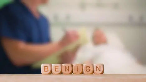 Medical Concept With Wooden Letter Cubes Or Dice Spelling Benign Against Background Of Nurse Talking To Patient In Hospital Bed