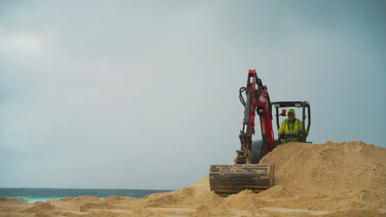 An orange construction excavator scoops and drops sand on a beach by the ocean mid shot