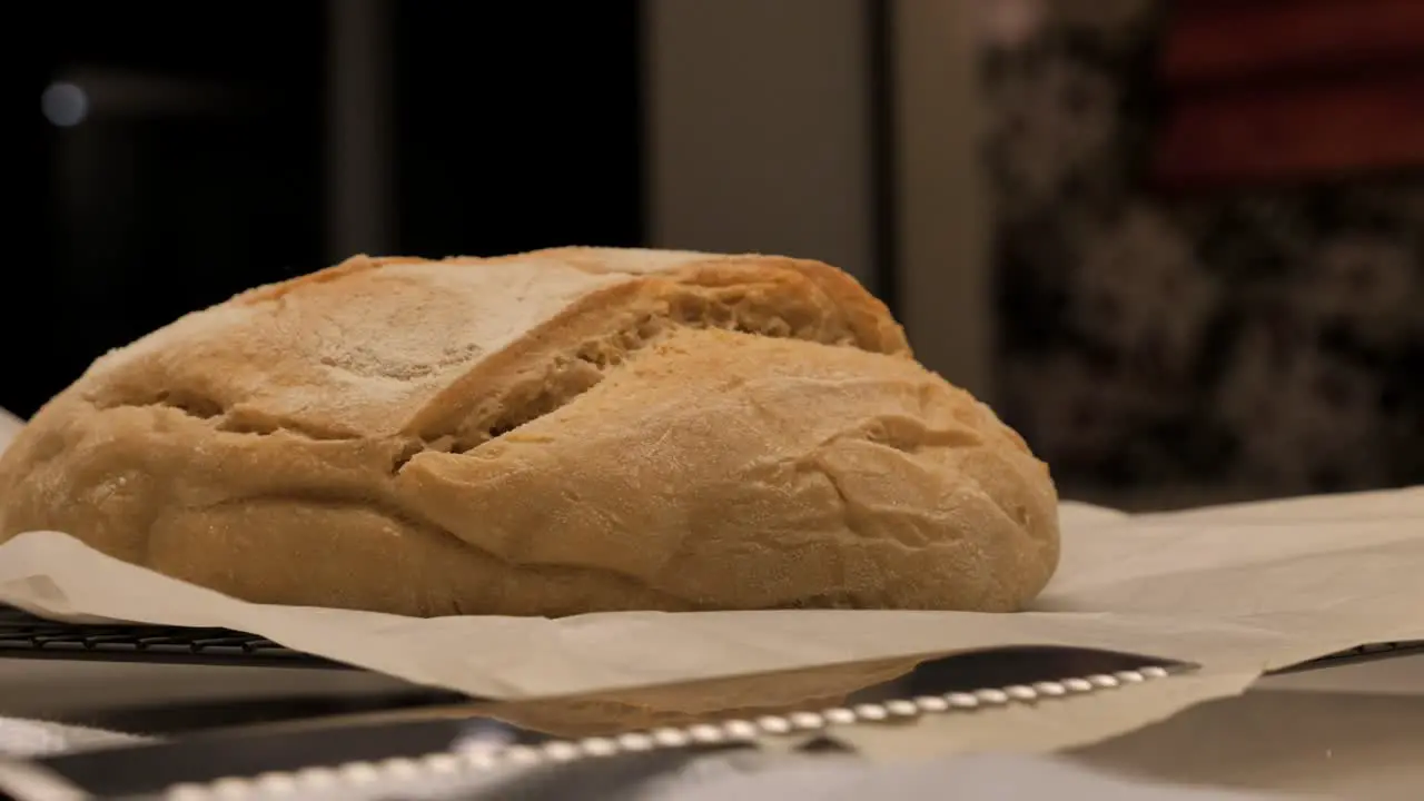 Slow pan around freshly baked loaf of sour dough bread topped with flour sitting on kitchen bench with tea towel and tray low depth of field