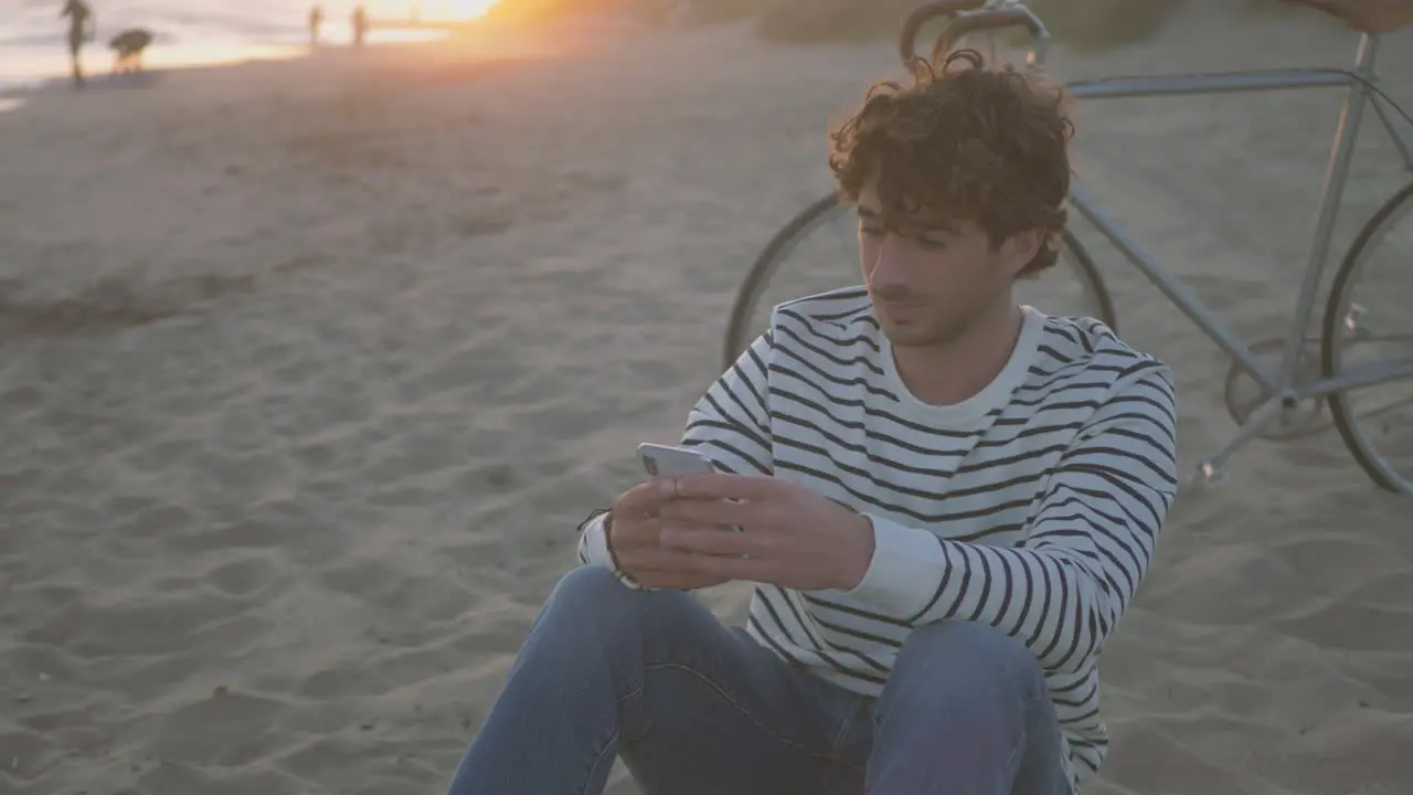 Man Sitting On The Sand At The Beach Using The Phone Next To His Bike