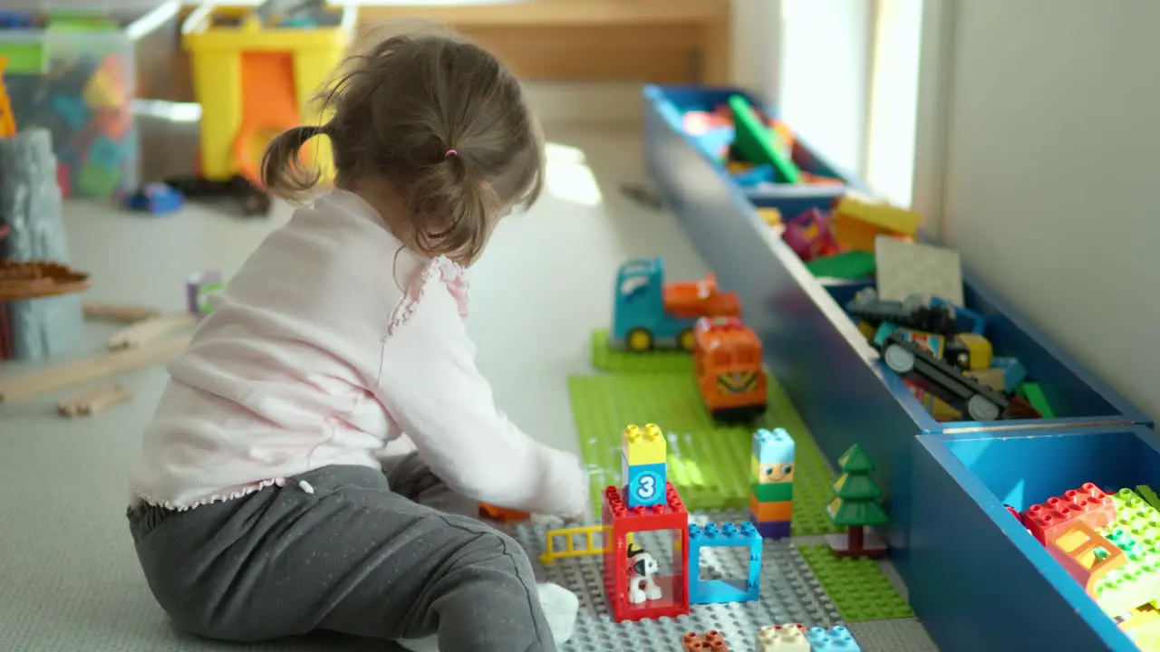 Cute funny little girl playing with construction toy blocks at home sitting on a floor by the window