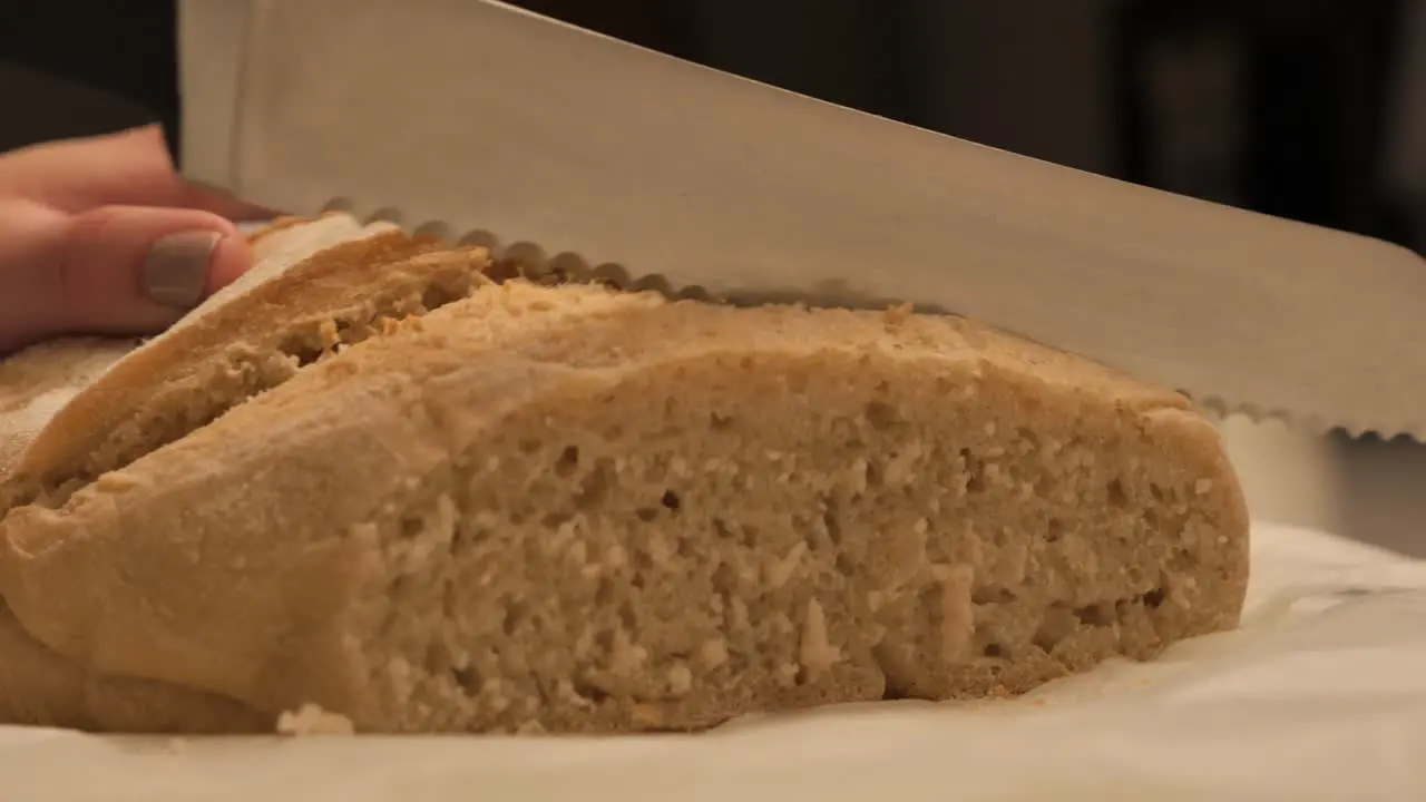 Female hand holding and cutting through freshly baked loaf of sour dough bread with sharp serated kitchen knife on benchtop