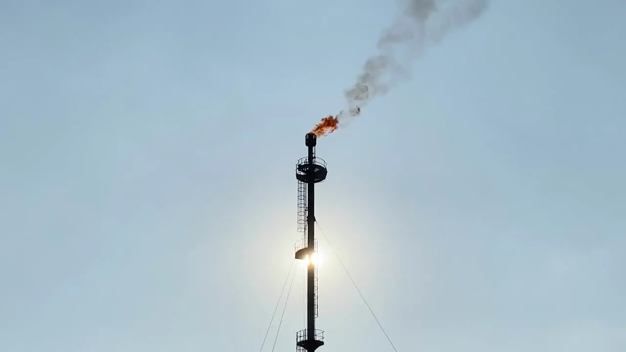 Flare stack refinery burning giant flame and smokestack sun behind clear sky