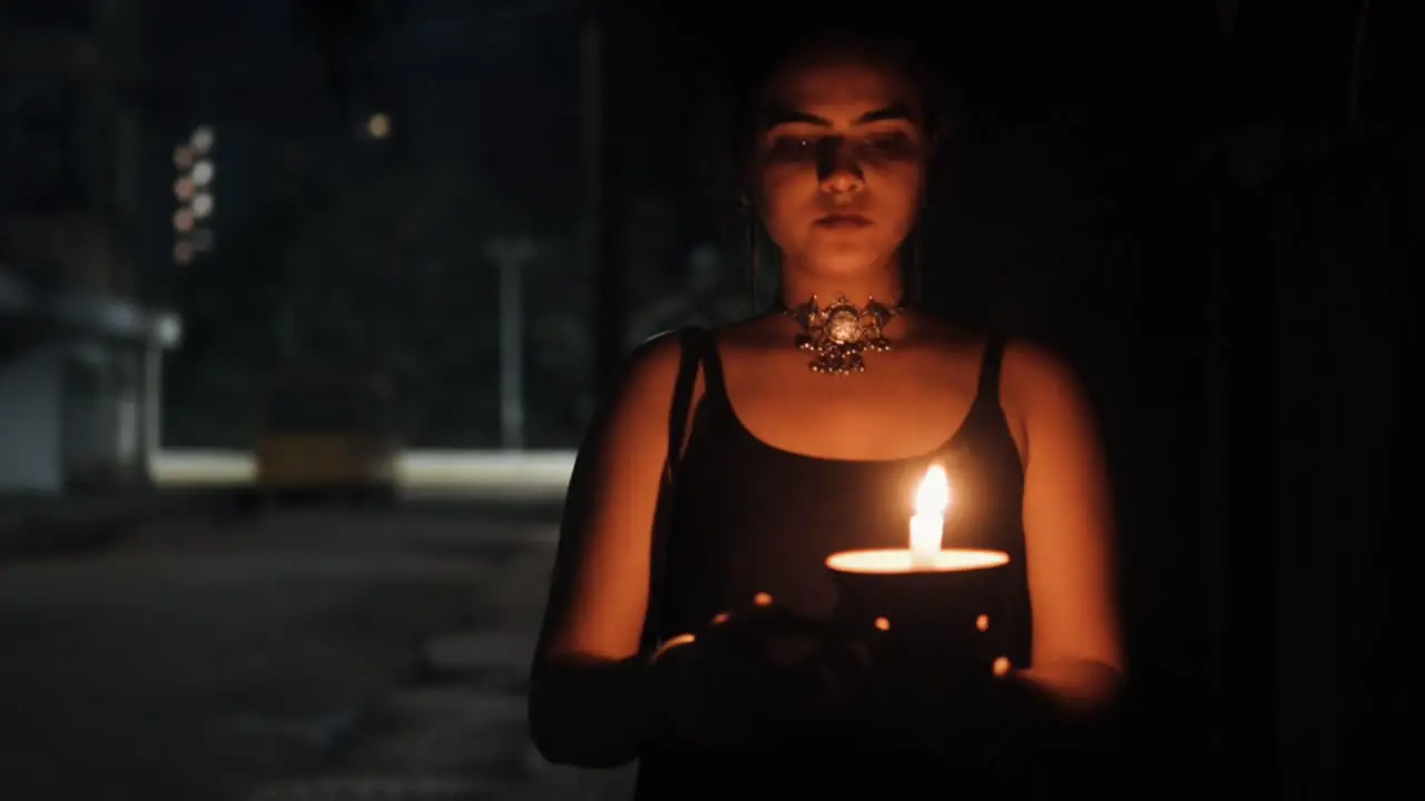 Front view of woman in black dress walking along dark street holding burning candle in hands with the background blur