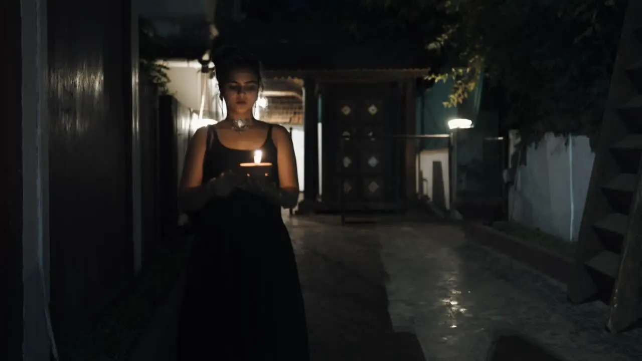 Front view of woman in black dress walking down dark hallway holding a burning candle in hands