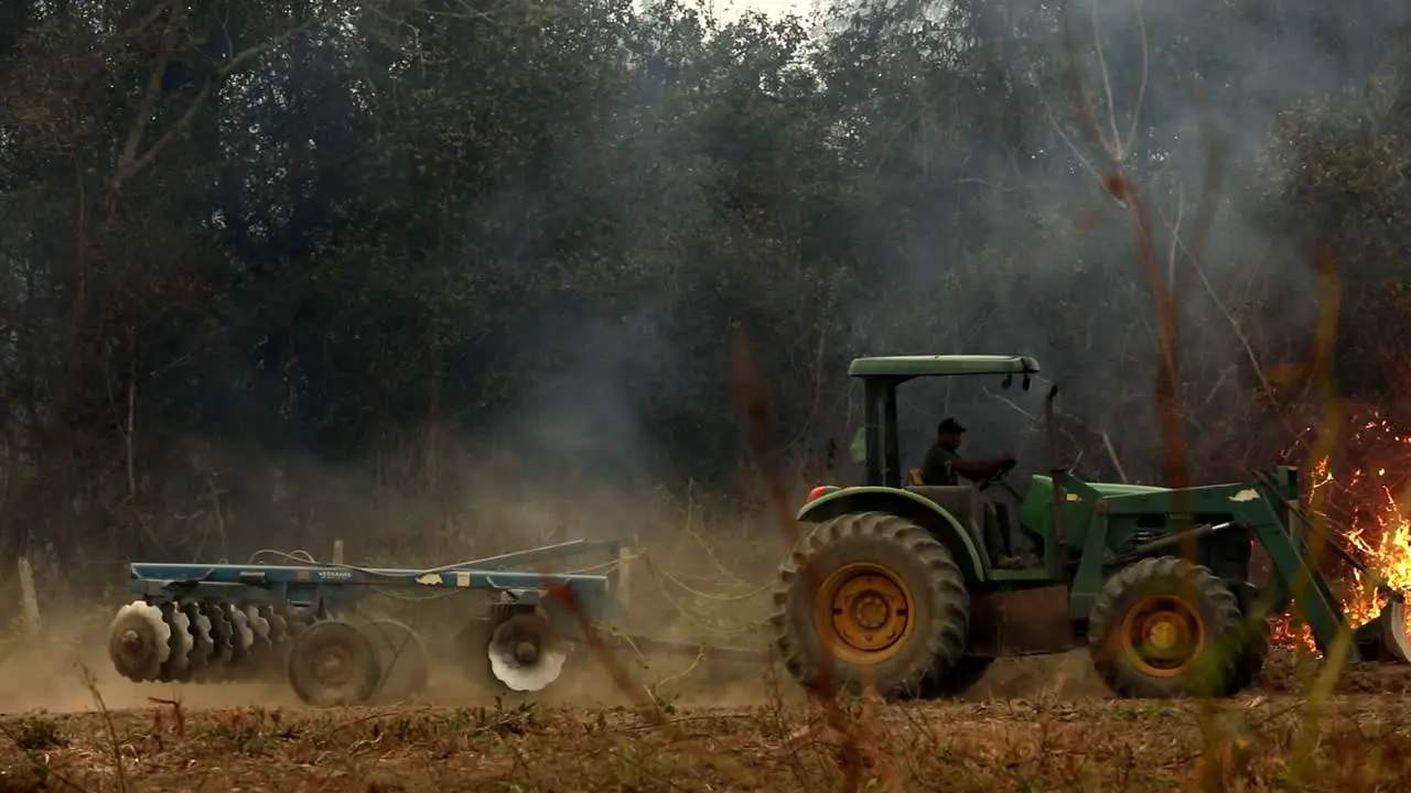 Tractor drives across a field with a wildefire burning brush in the background