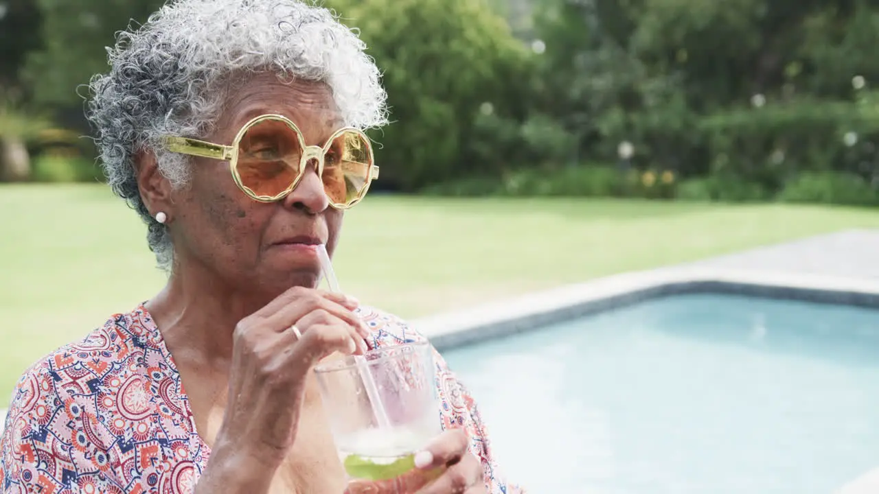 Happy senior african american woman in sunglasses by pool drinking cocktail copy space slow motion