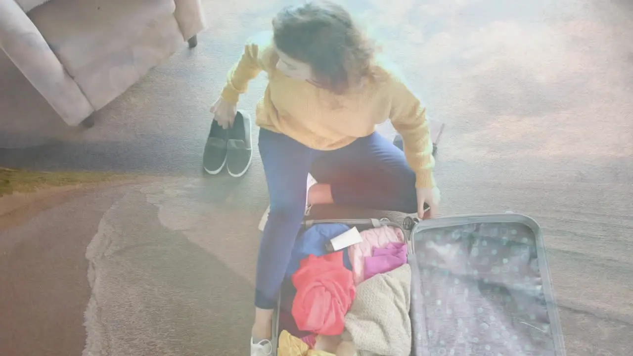 Animation of happy caucasian woman sitting on floor packing suitcase over beach landscape