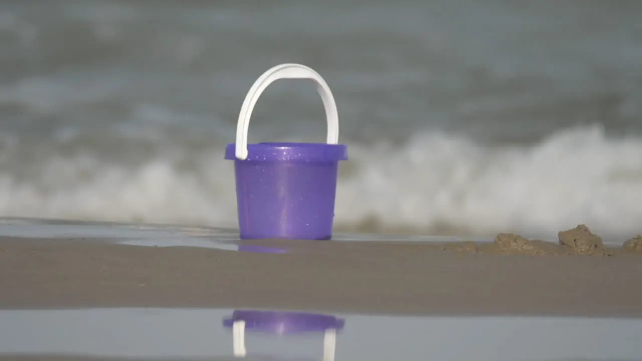 A purple toy bucket on the sand on the beach with sparkling waves on the background