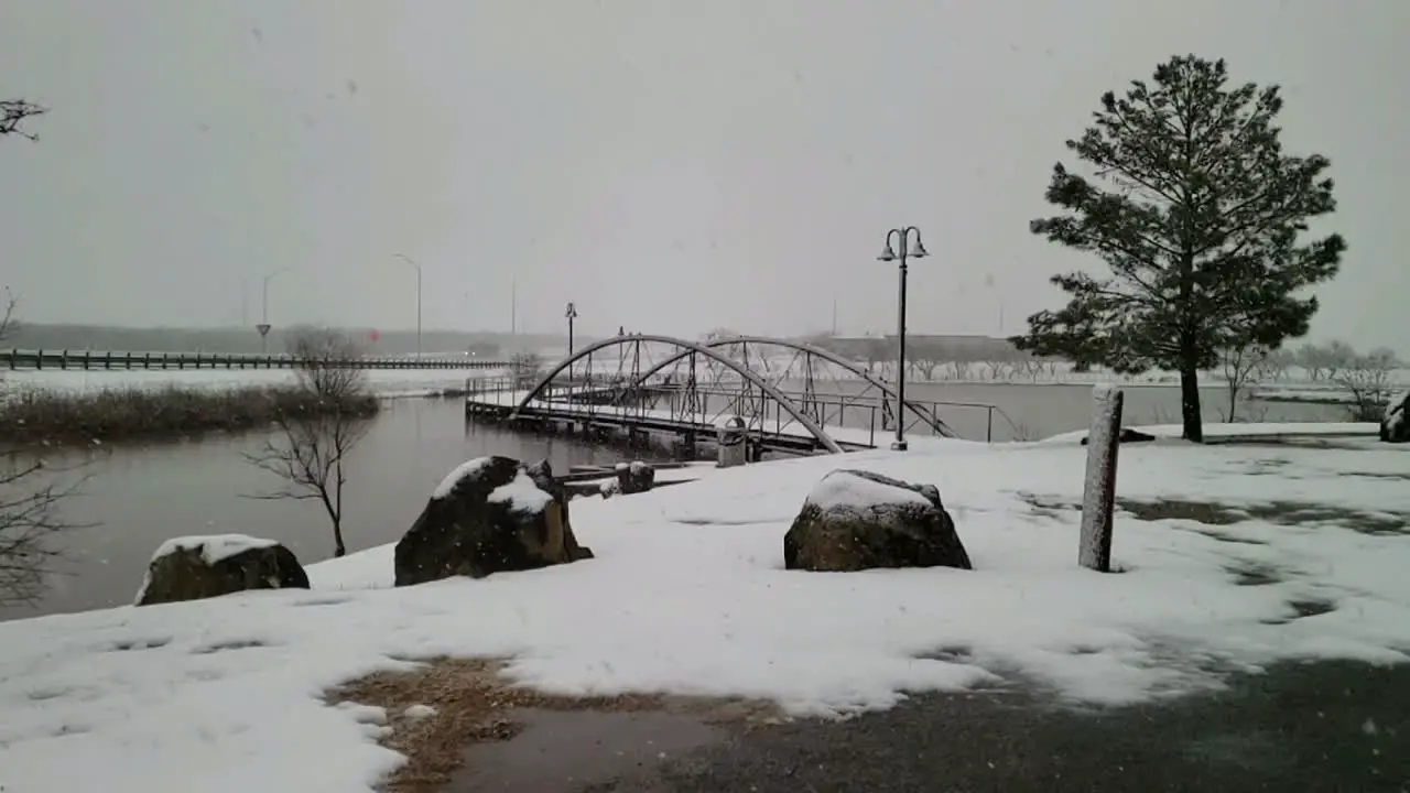 Watching the snow fall in slow motion on small Texas city lake with beautiful walking bridge trees lake rocks and gray skies