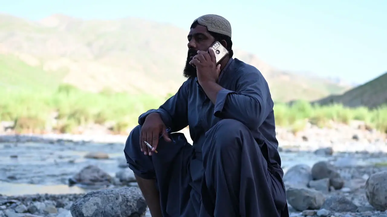 Pakistani Muslim Male Sat Beside River Talking On Mobile Phone With Cigarette In Hand In Sindh