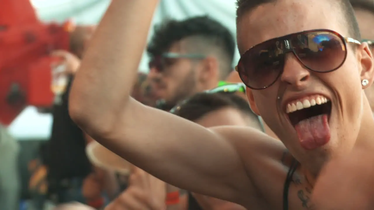 Spanish looking male with chest tattoos and tinted sunglasses cheers at a music festival with his tongue out and waves his arms