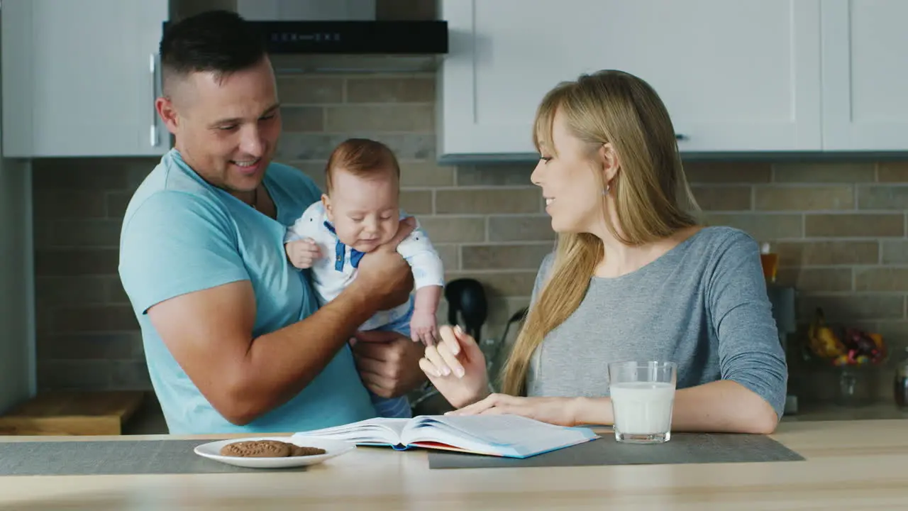 Young Family In The Kitchen Mom Reading A Book With The Baby Daddy Is Near