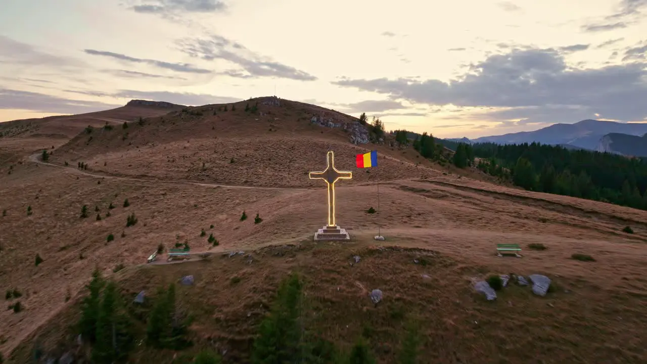 Aerial backwards shot of lighting cross with waving Romanian flag on summit of Mount Dichiu at sunset