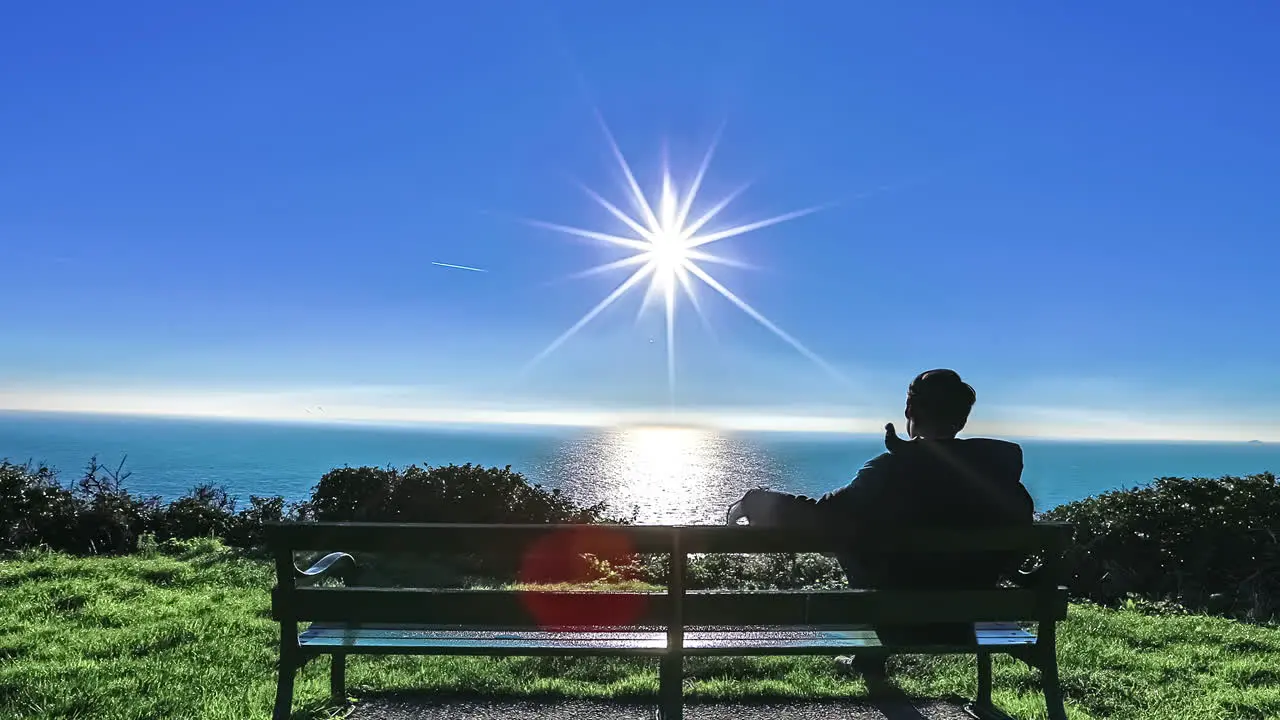 Young man on a park bench watching the ocean with the sun reflecting off the water siding time lapse