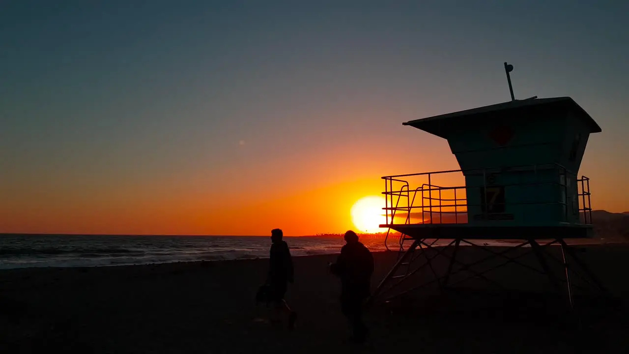 Slow and low sideways sunset shot of Lifeguard house  tower with silhouettes of people passing in front at sunset at San Buenaventura State Beach in Ventura California United States